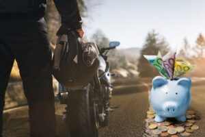 Una moto e una persona in piedi con il casco, a destra un salvadanaio con banconote e monete